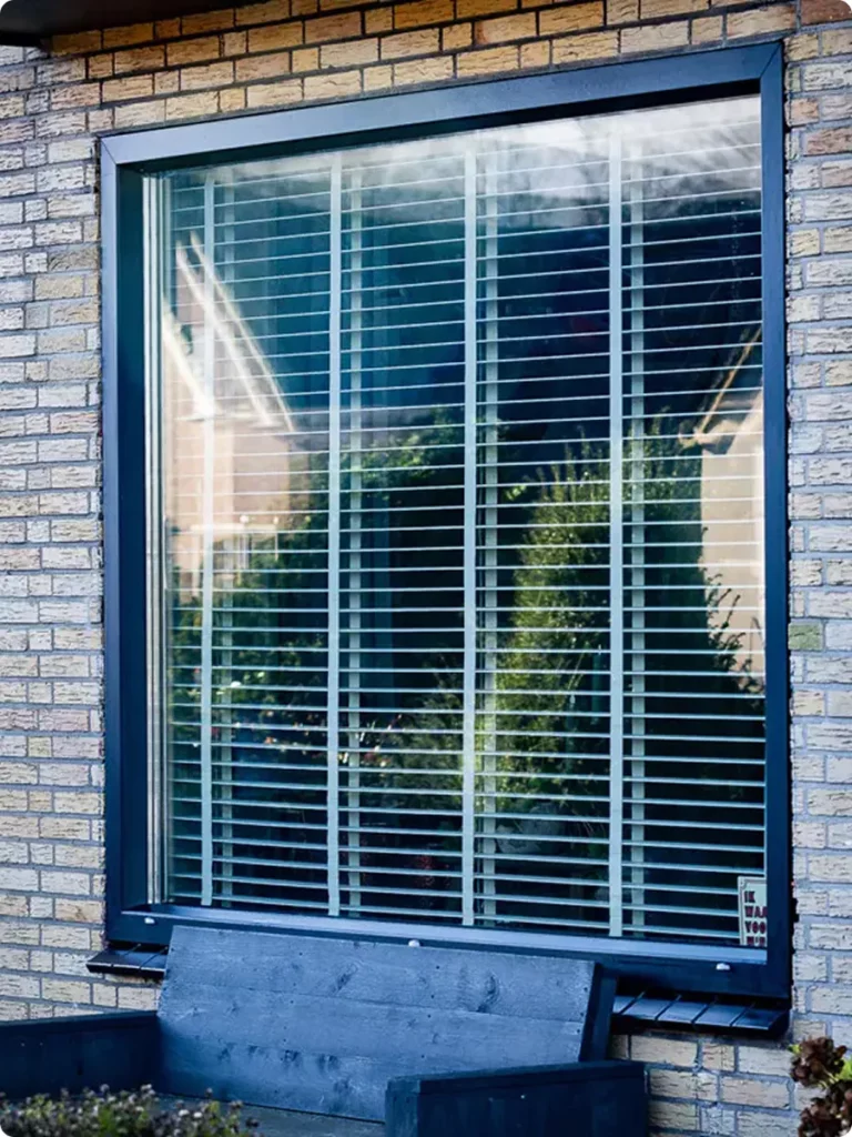 Groot raam met zwart aluminium kozijn en horizontale jaloezieën in een bakstenen gevel.